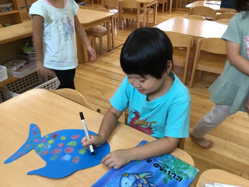 虹色の魚の絵本を先生に読んでもらった子どもたち。
鱗をもらったお友だちの魚のように、自分たちもキラキラ光る鱗を見てみたい！
そんな思いに先生がいろんな色の砂を用意してくれました。
砂が鱗に見えるように、慎重に魚の台紙に乗せていく表情はとても真剣。
集中力はこういった経験から培われるのかもしれません。
出来上がった魚をお友だち同士で褒め合う姿は絵本から何かを感じ取った後に
作ったからかもしれませんね。