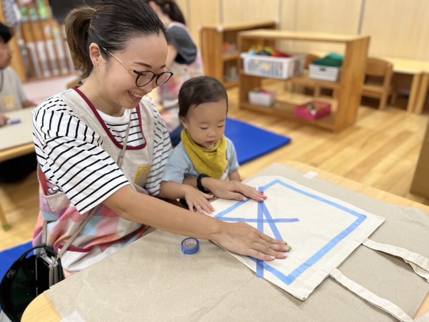 今週は1歳児さんの保育参観があります。<br />
保護者の方にもぽけっとランドのエプロンを着用して参加して頂きました。<br />
テープやシールを剝がしたり貼ったりすることが大好きな1歳児さん。<br />
今日は布製のバックにお母さんやお父さんと一緒にマスキングテープを貼ります♪